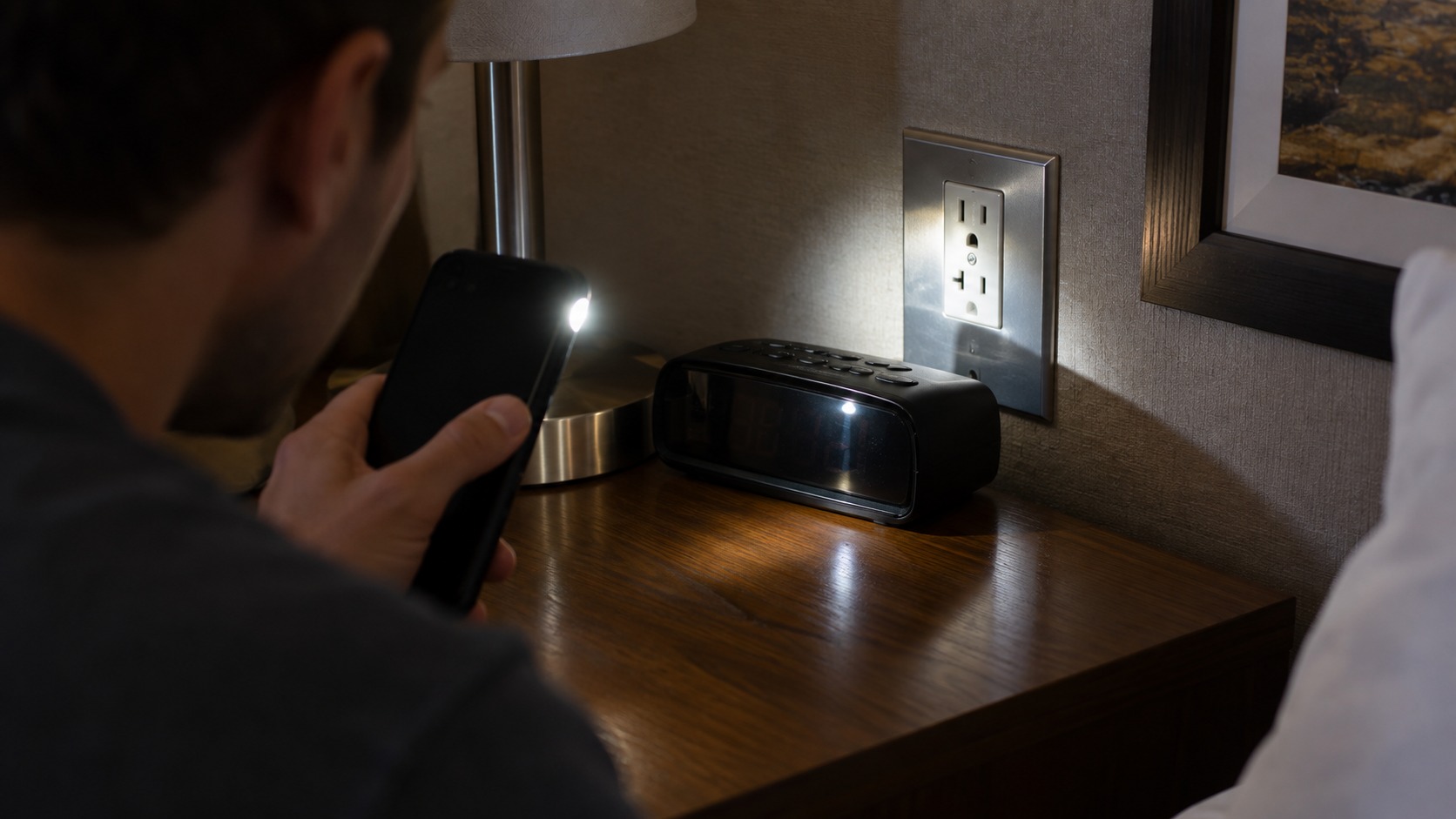 Phone flashlight sweeping across a hotel alarm clock and outlet to check small reflections at close range.