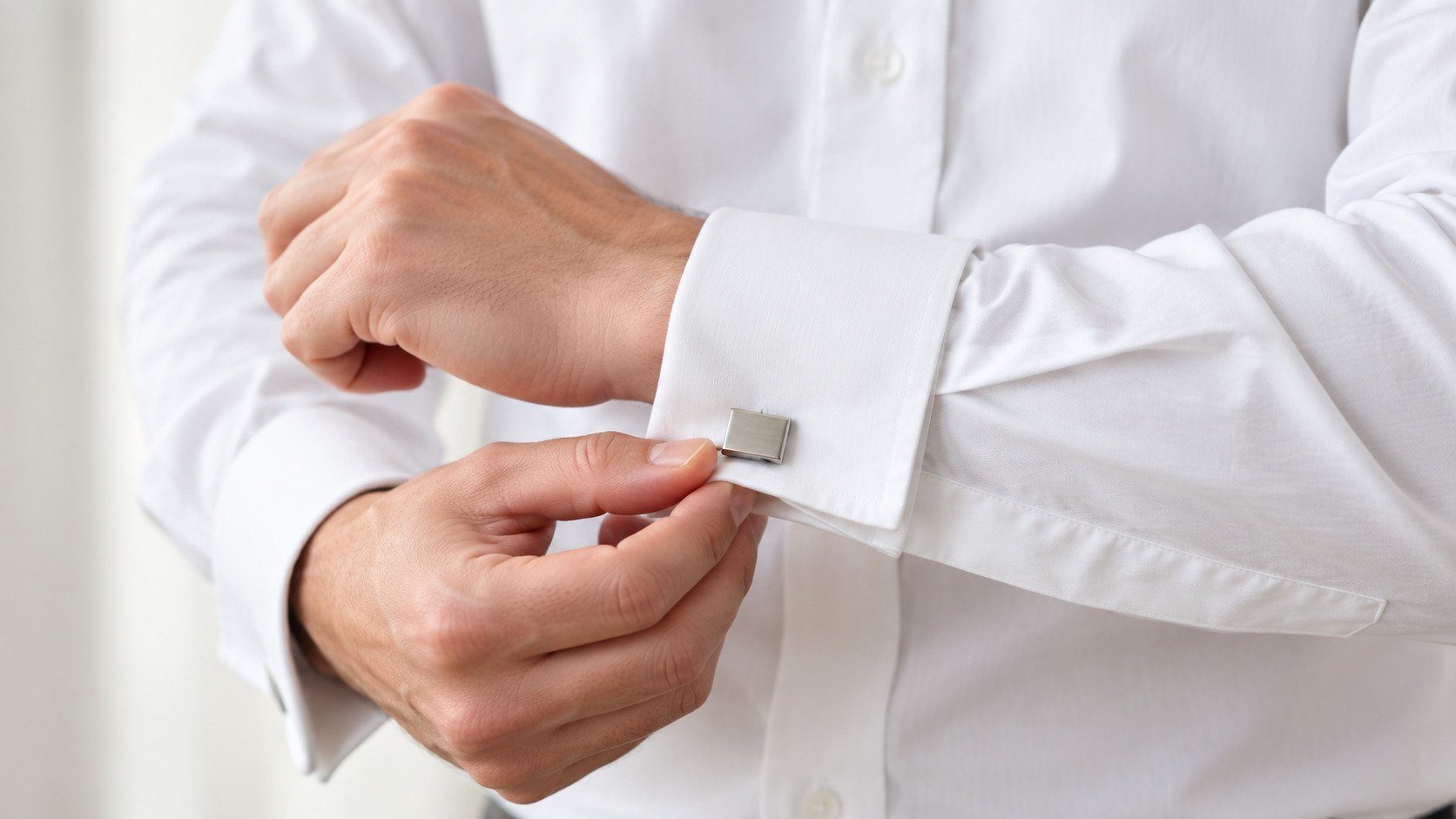 Hands folding the extended double cuff of a white formal shirt and fastening it with a small plain silver cufflink.